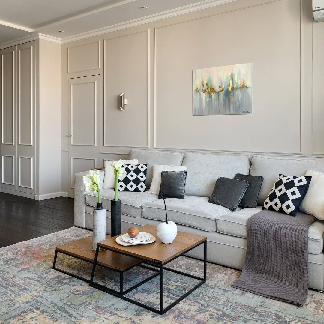 Elegancki salon w beżach: sofa z poduszkami, stolik kawowy z dekoracjami, obraz na ścianie z panelami. Nowoczesne wykończenie wnętrza z dbałością o detale.