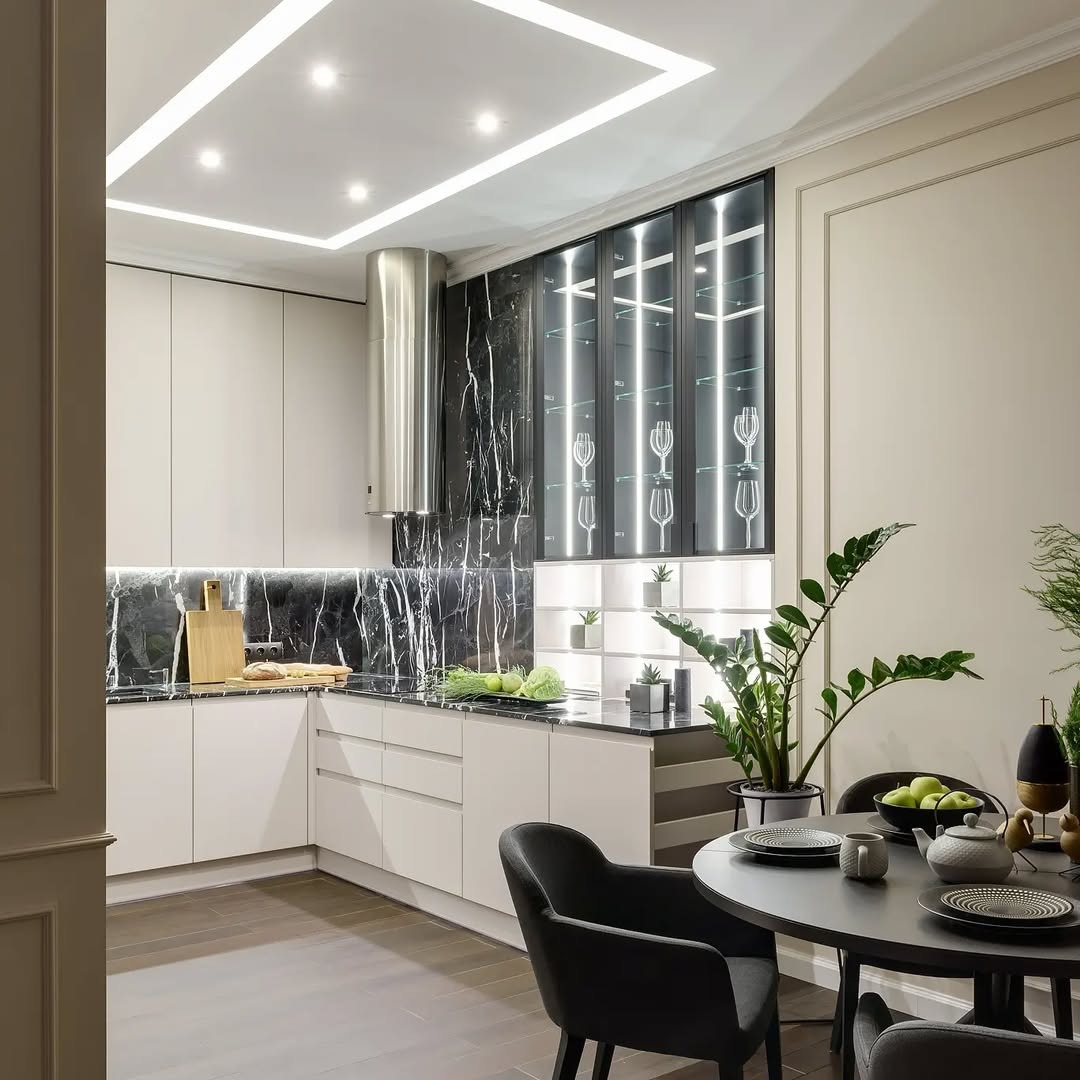 Nowoczesna kuchnia z czarnym marmurem na ścianie, białymi szafkami i oświetleniem LED. Na stole czarne krzesła i dekoracje roślinne. Eleganckie wykończenie wnętrza.