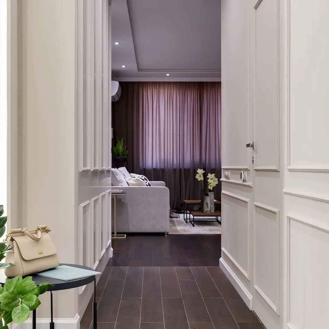 Eleganckie wnętrze: ciemna podłoga, kremowe ściany z dekoracyjnymi listwami, widok na salon z sofą i zasłonami. Torebka na stoliku w pierwszym planie.