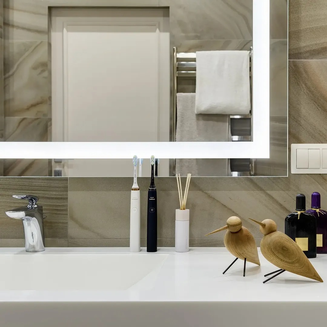 Nowoczesna łazienka z podświetlanym lustrem, chromowaną baterią umywalkową i dekoracyjnymi figurkami ptaków na blacie. Stonowana kolorystyka płytek i akcesoriów.