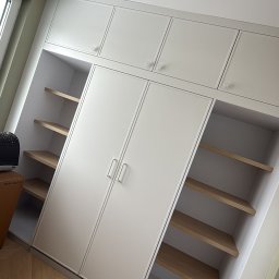 Konrad Nizioł - Biała szafa wnękowa z drewnianymi półkami, widok z dołu, parkiet w jodełkę na podłodze. Minimalistyczny design, jasne wnętrze, nowoczesne wykończenie.