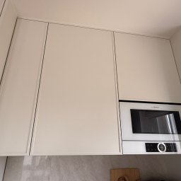Konrad Nizioł - Jasne, minimalistyczne szafki kuchenne pod sufitem z wbudowaną mikrofalówką Bosch. Ujęcie z dołu, widoczny fragment blatu i ściany.