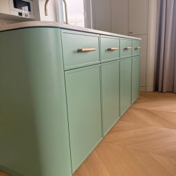 Konrad Nizioł - Pastelowa, pistacjowa wyspa kuchenna z drewnianymi uchwytami na tle parkietu w jodełkę i zasłony. Minimalistyczny design z zaokrąglonymi krawędziami.