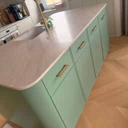 Konrad Nizioł - Nowoczesna wyspa kuchenna w pastelowej zieleni z marmurowym blatem i złotymi uchwytami, na tle parkietu w jodełkę. Minimalistyczny design z elementami luksusu.