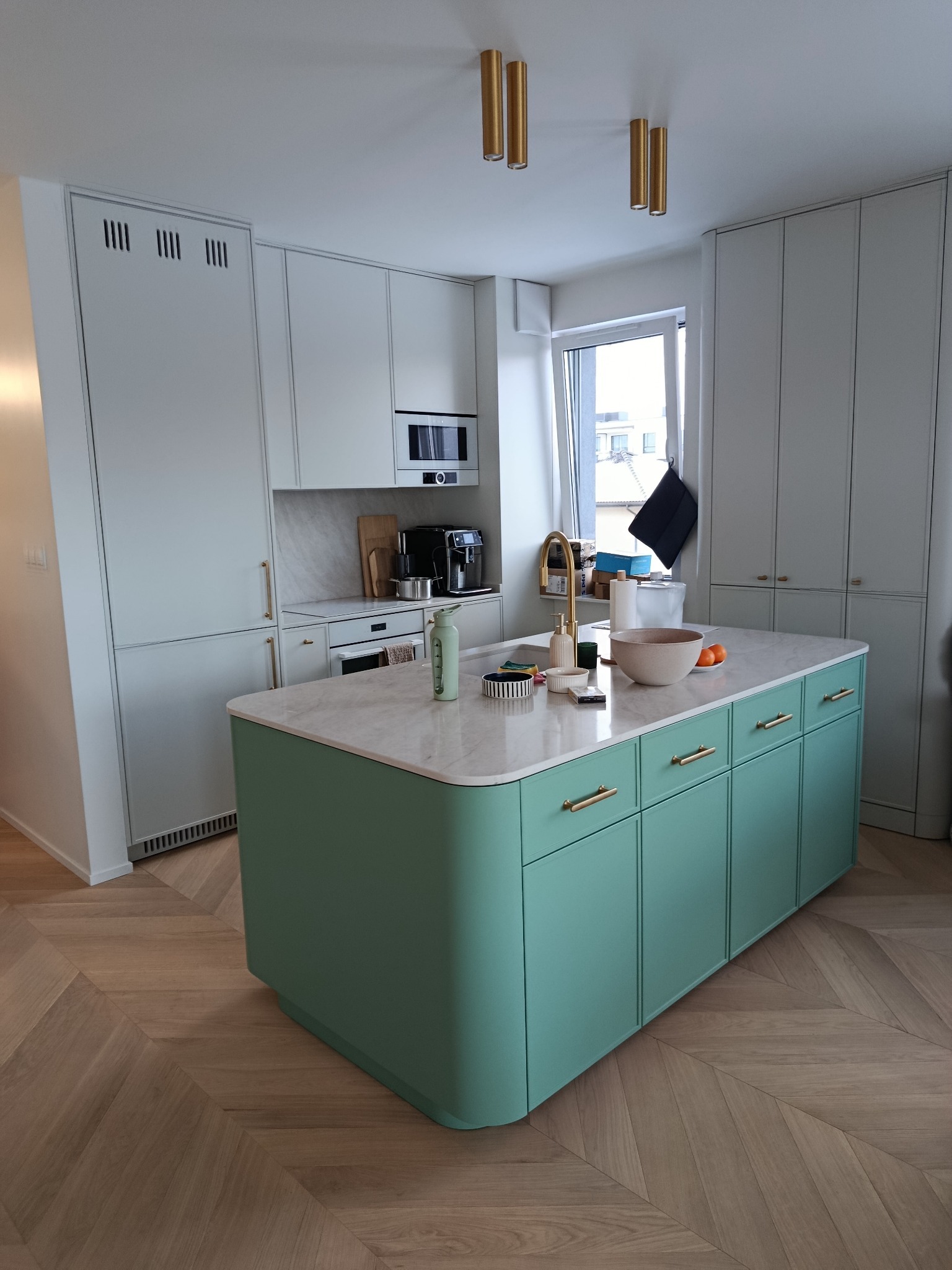 Nowoczesna kuchnia z wyspą w odcieniach bieli i mięty, złote akcenty, parkiet w jodełkę. Minimalistyczny design, naturalne światło, marmurowy blat wyspy.