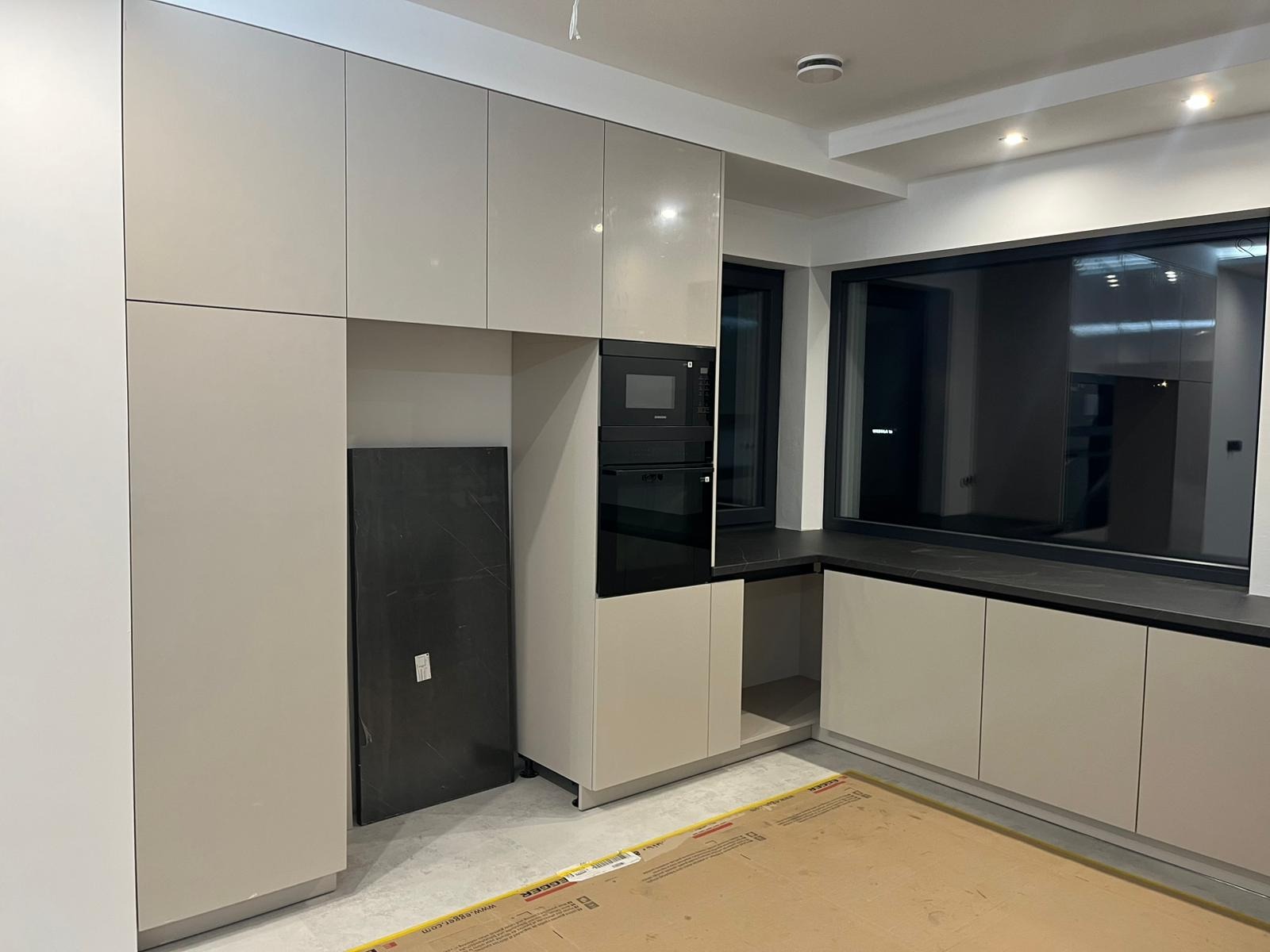 Jasna, nowoczesna kuchnia z zabudową pod sufit, czarnym blatem i dużym oknem. Widoczny fragment płyty meblowej oparty o ścianę. Minimalistyczny design.