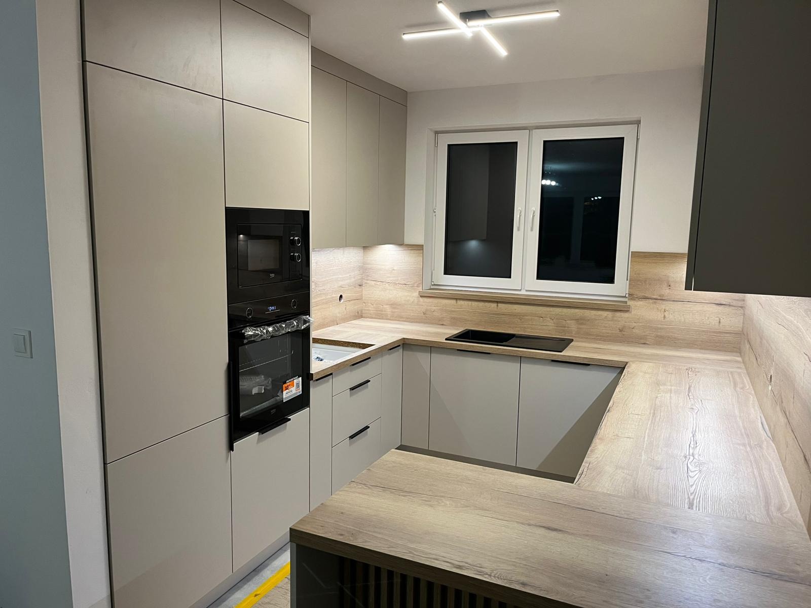 Jasna kuchnia z drewnianym blatem i czarnym sprzętem AGD. Ujęcie z bliska prezentuje nowoczesny design i funkcjonalne rozwiązania w zabudowie kuchennej.