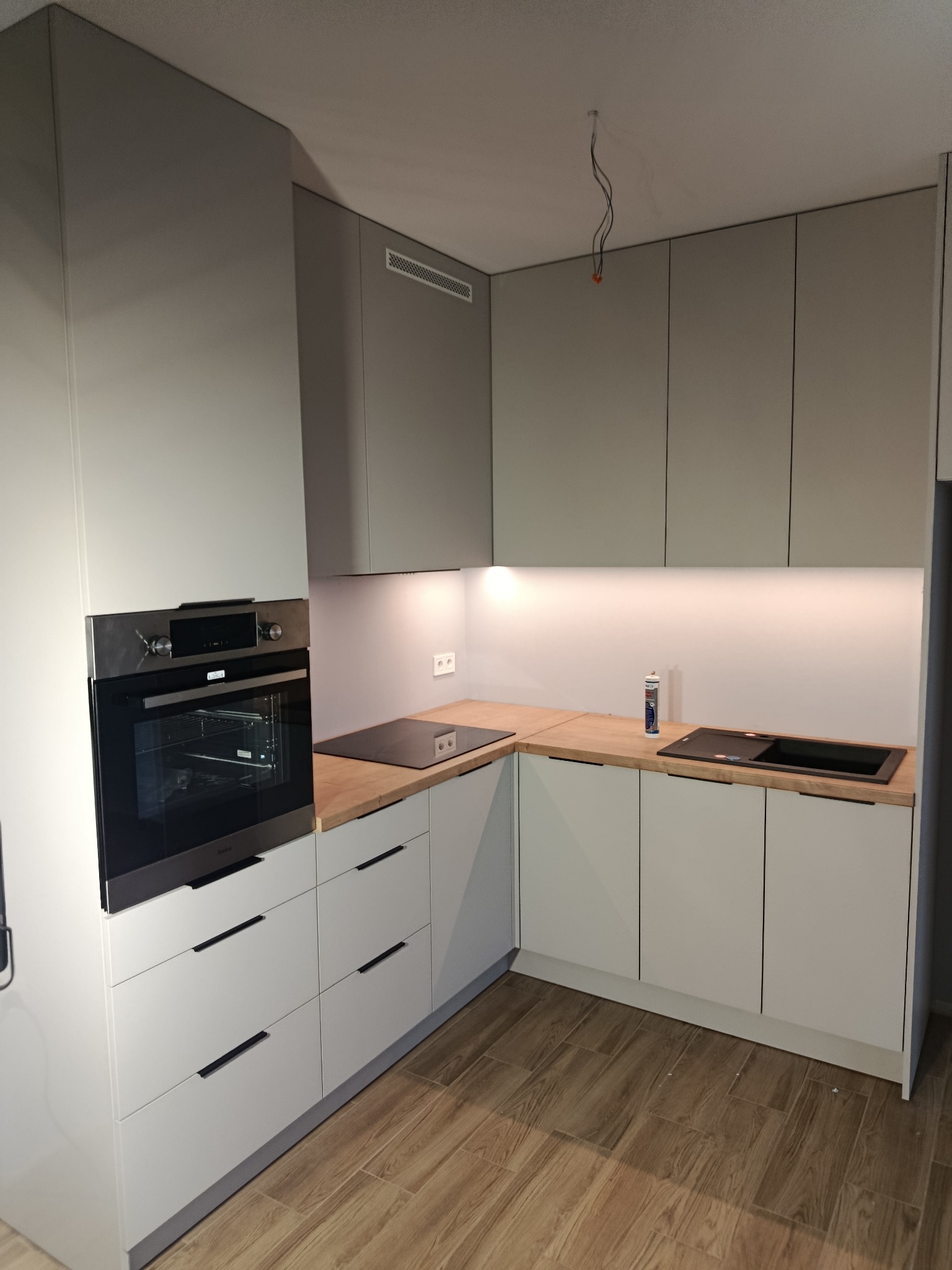 Nowoczesna, jasnoszara kuchnia z drewnianym blatem, czarną płytą indukcyjną i piekarnikiem Amica. Minimalistyczne uchwyty, oświetlenie LED pod szafkami.