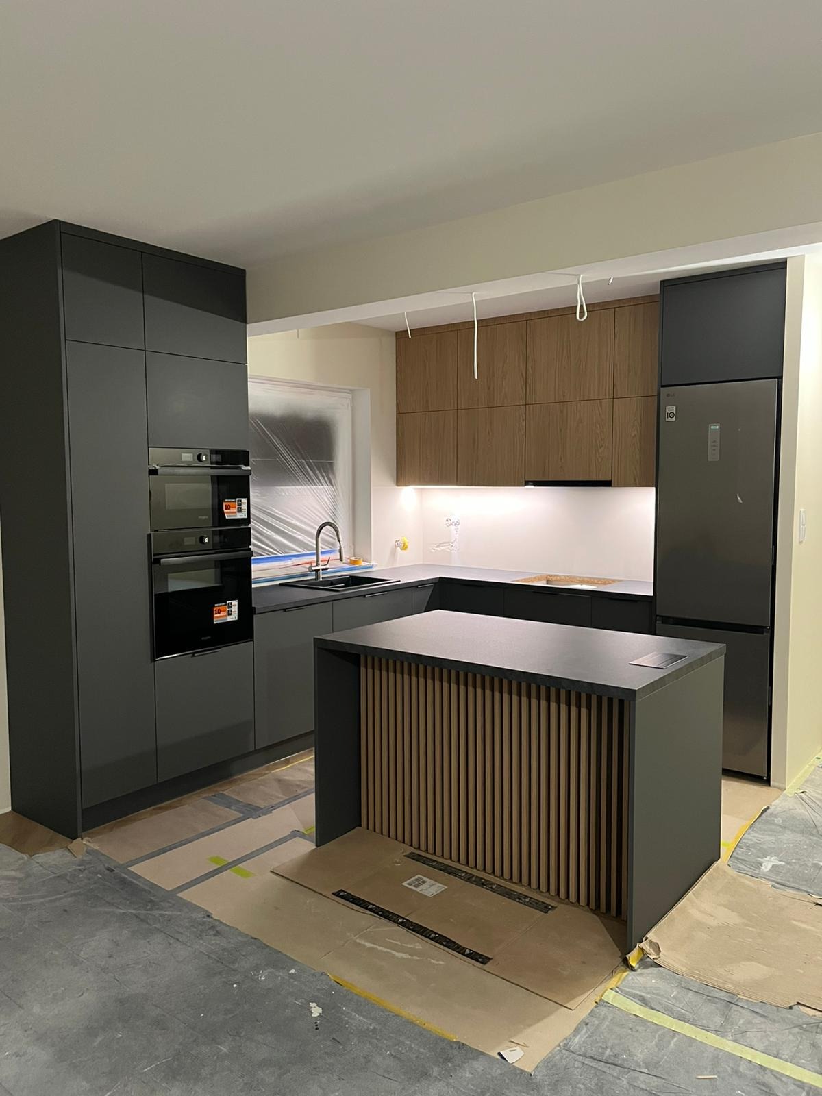 Nowoczesna kuchnia z szarymi szafkami, drewnianymi akcentami wyspy i sprzętem AGD w zabudowie. Okno zasłonięte folią ochronną. Podłoga zabezpieczona kartonem.