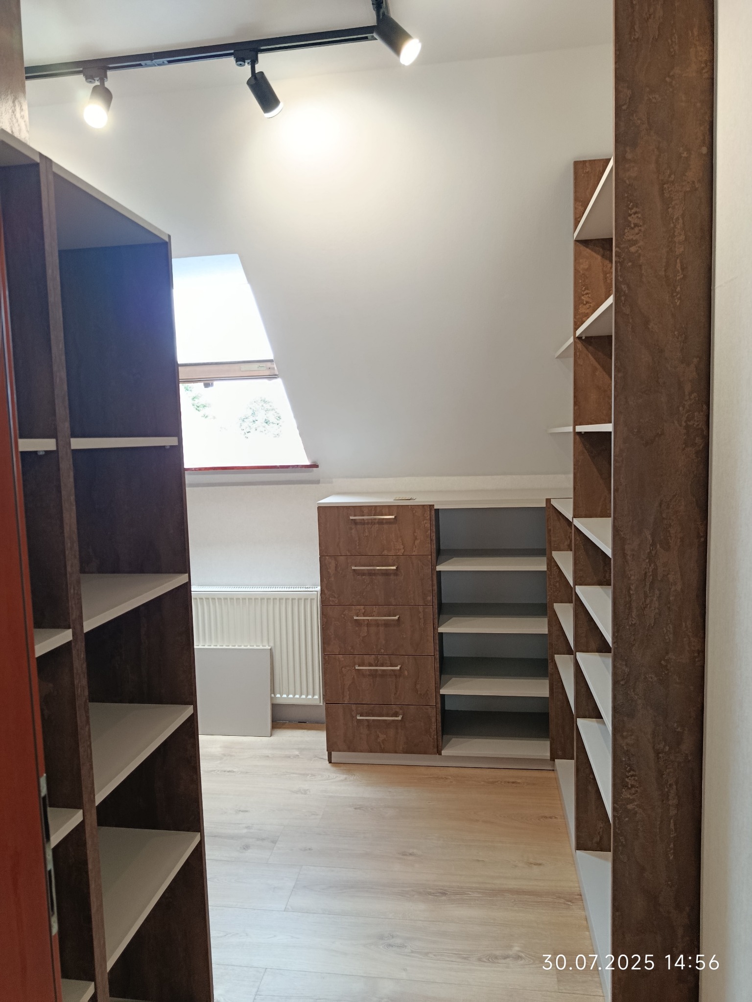 Szafa wnękowa z szufladami i półkami w brązowym odcieniu, dopasowana do skosu dachu. Realizacja w Lublinie, oświetlenie punktowe na suficie.