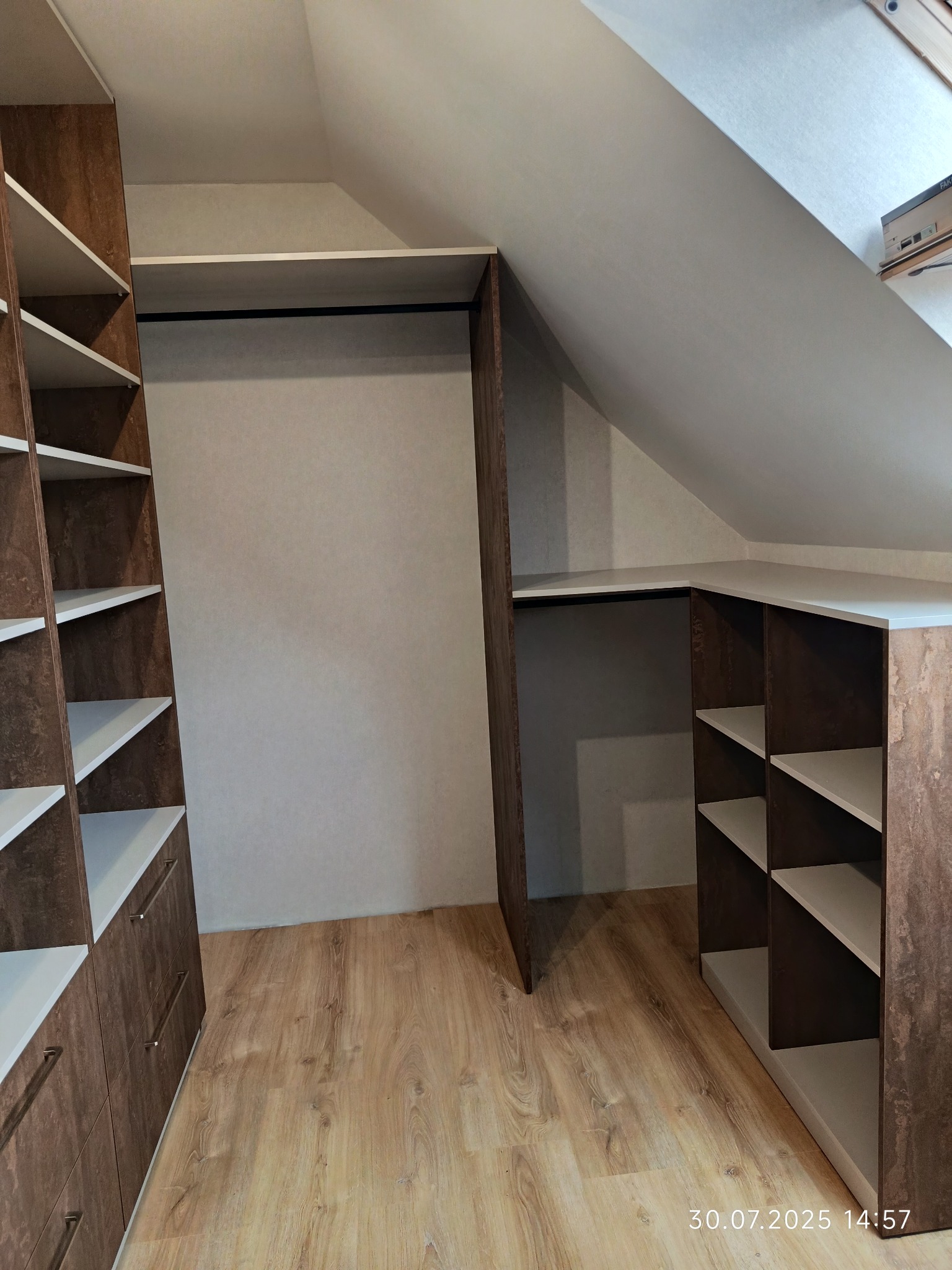 Szafa wnękowa z brązowymi frontami i białymi półkami, dopasowana do skosu poddasza. Widoczne szuflady i otwarte półki. Jasna podłoga i ściany.