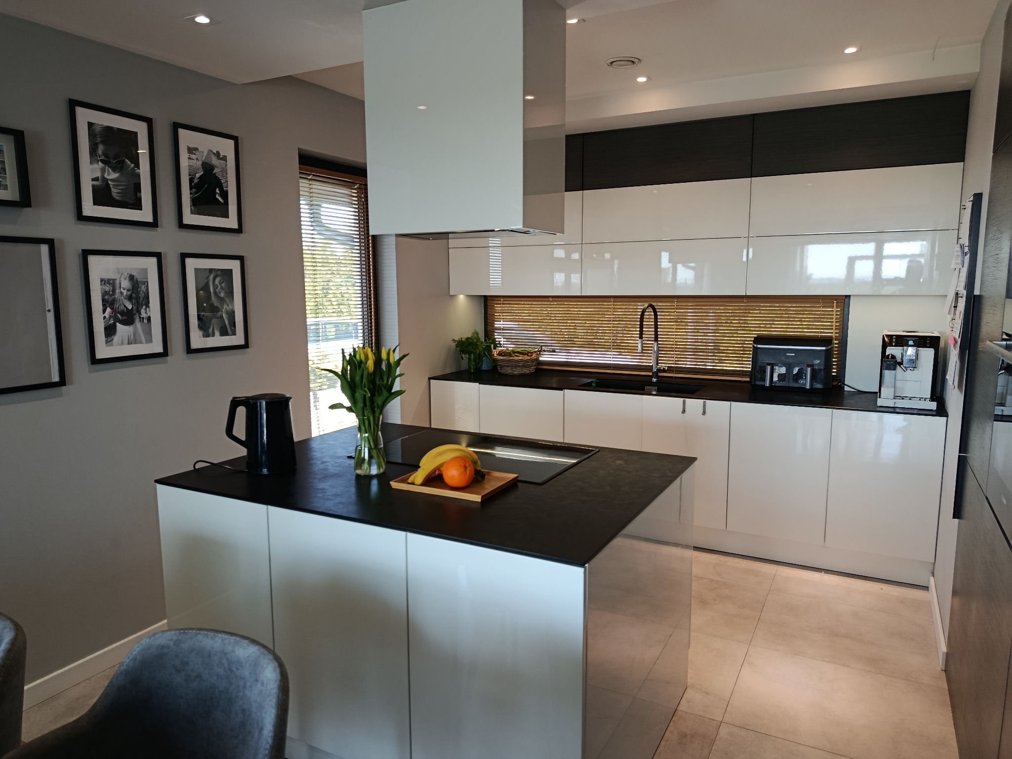 Nowoczesna kuchnia z wyspą: białe, lakierowane fronty, czarny blat, minimalistyczny okap. Na wyspie owoce i kwiaty. W tle okno z żaluzjami.