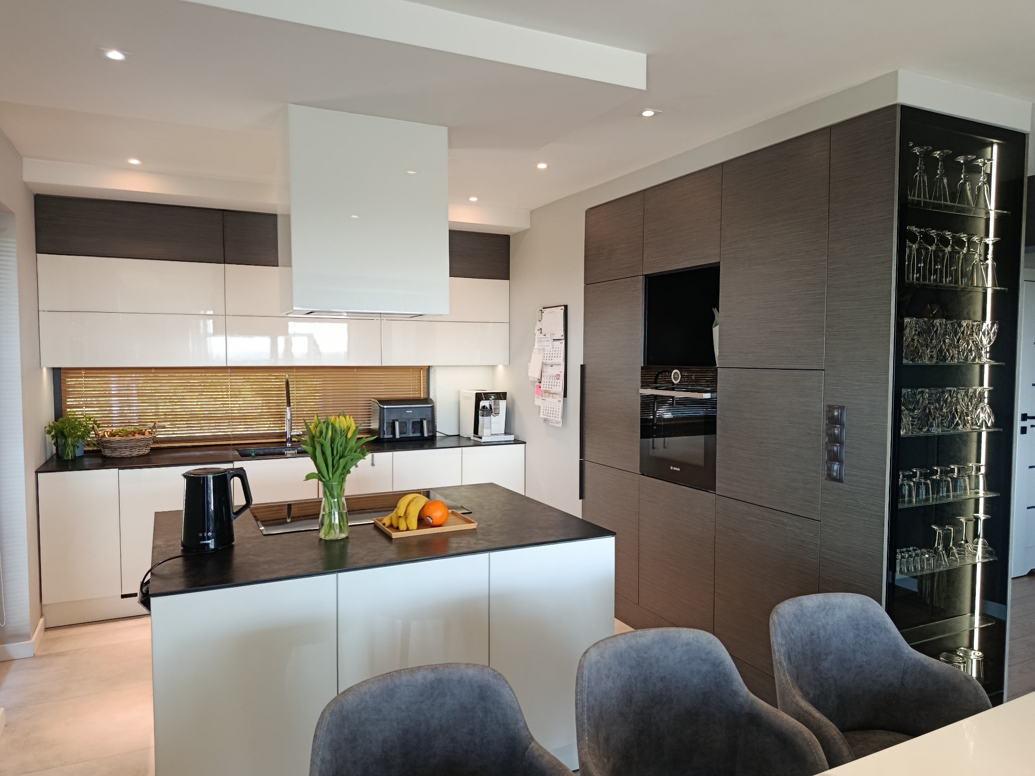 Nowoczesna kuchnia z wyspą, fronty białe i w odcieniach brązu. Na wyspie czajnik, kwiaty i owoce. W tle zabudowa z piekarnikiem i przeszklona witryna z kieliszkami.