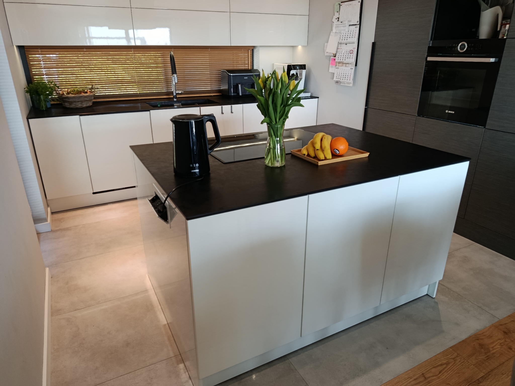 Nowoczesna kuchnia na wymiar z wyspą w Lublinie. Białe fronty szafek, czarny blat, kwiaty w wazonie i owoce na wyspie. Minimalistyczny design, eleganckie wykończenie.