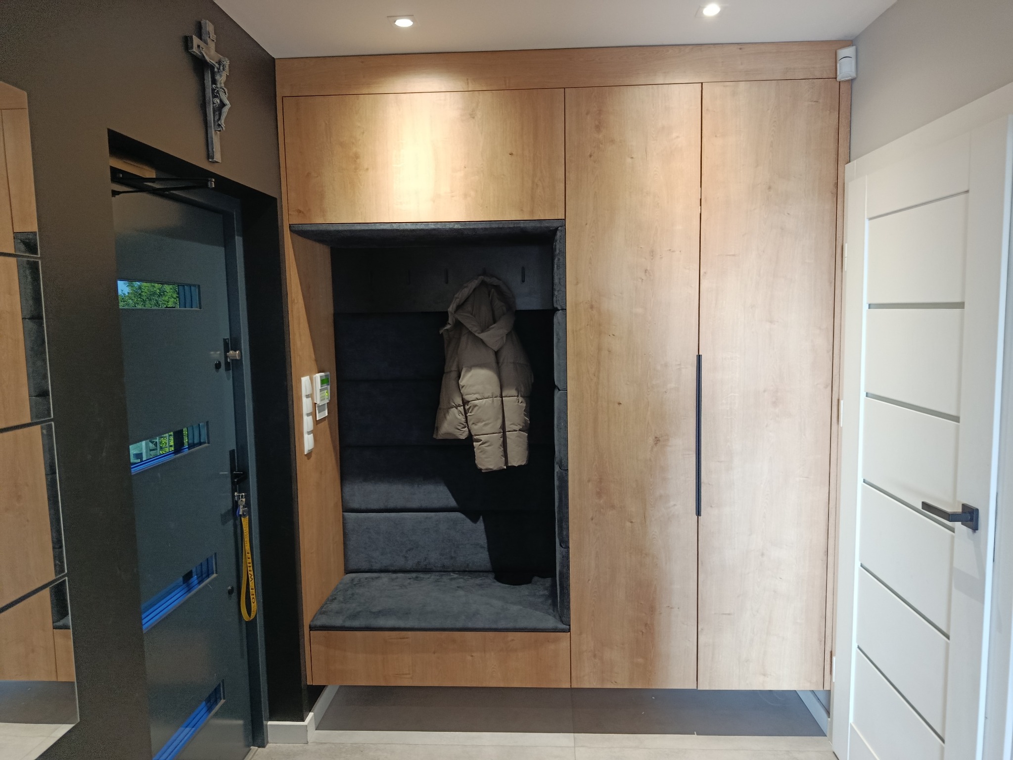 Drewniana szafa wnękowa z siedziskiem i wieszakiem na kurtkę, wkomponowana w przedpokój. Obok drzwi wejściowe i wewnętrzne. Na ścianie krucyfiks.