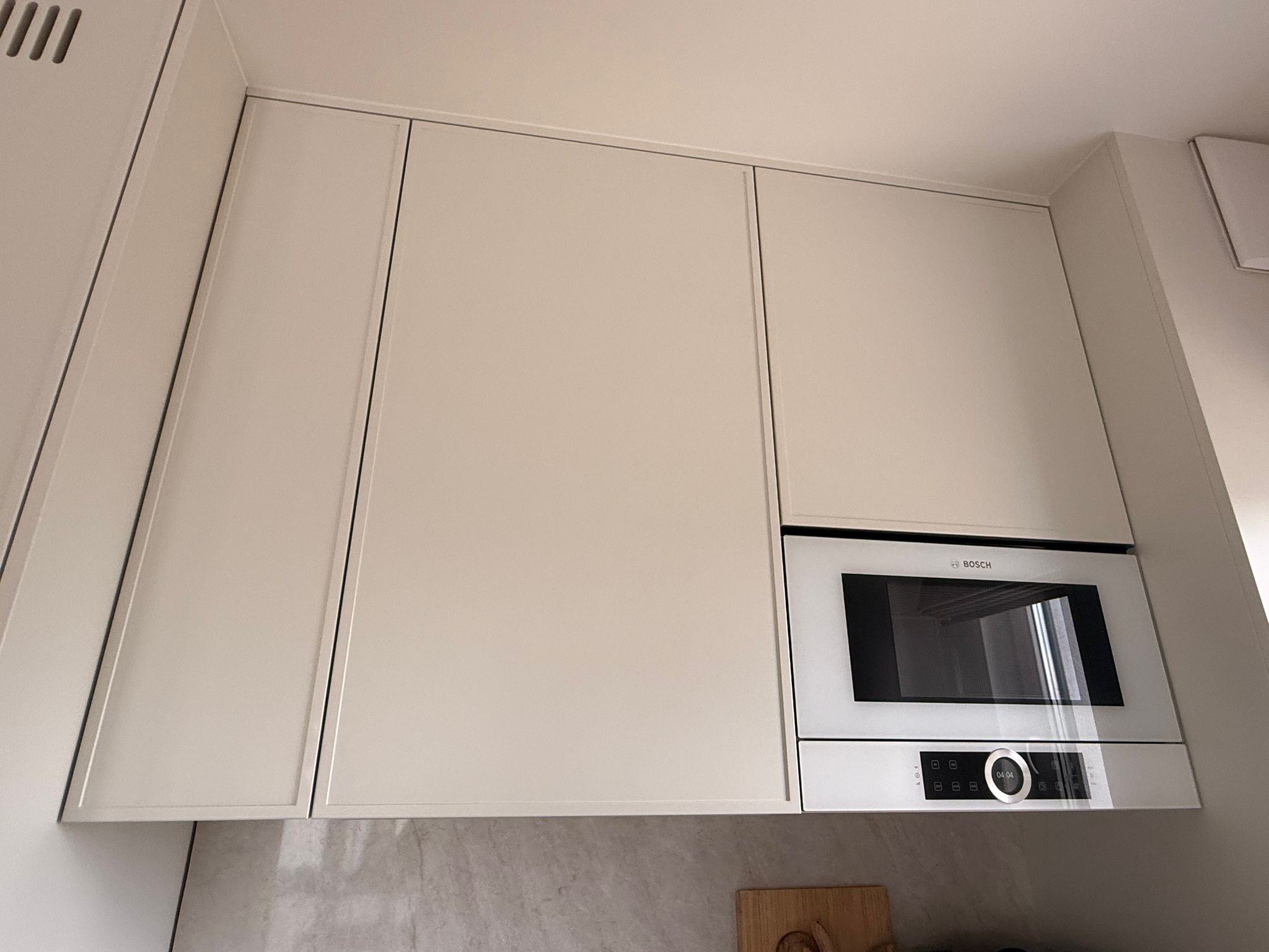 Jasne, minimalistyczne szafki kuchenne pod sufitem z wbudowaną mikrofalówką Bosch. Ujęcie z dołu, widoczny fragment blatu i ściany.