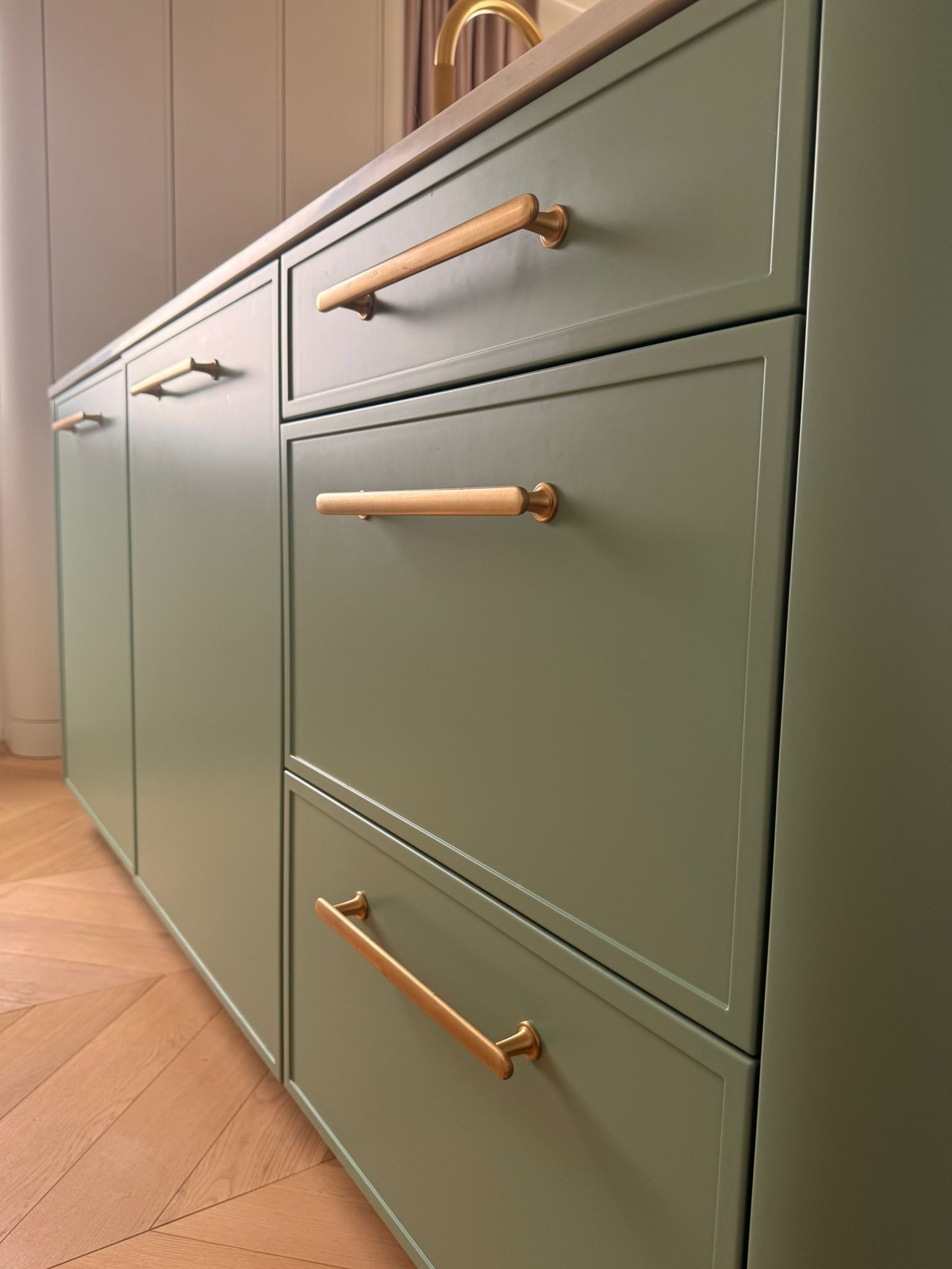 Zielona zabudowa kuchenna z drewnianymi uchwytami na tle parkietu w jodełkę. Ujęcie z dołu podkreśla nowoczesny design i dbałość o detale.