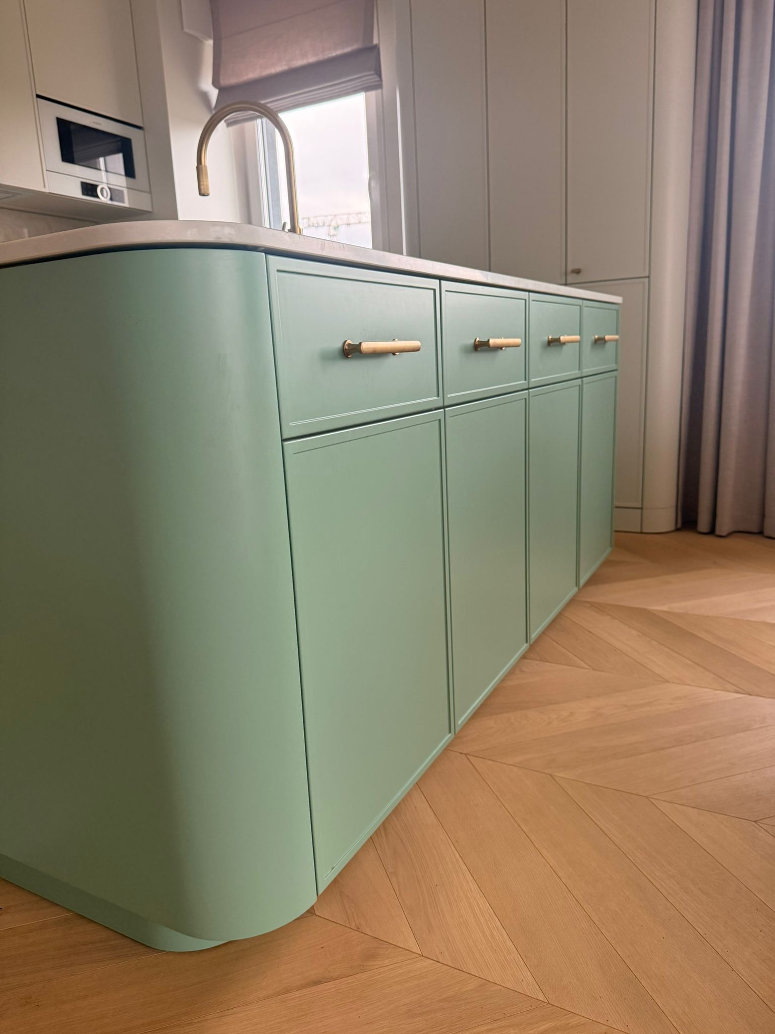 Pastelowa, pistacjowa wyspa kuchenna z drewnianymi uchwytami na tle parkietu w jodełkę i zasłony. Minimalistyczny design z zaokrąglonymi krawędziami.