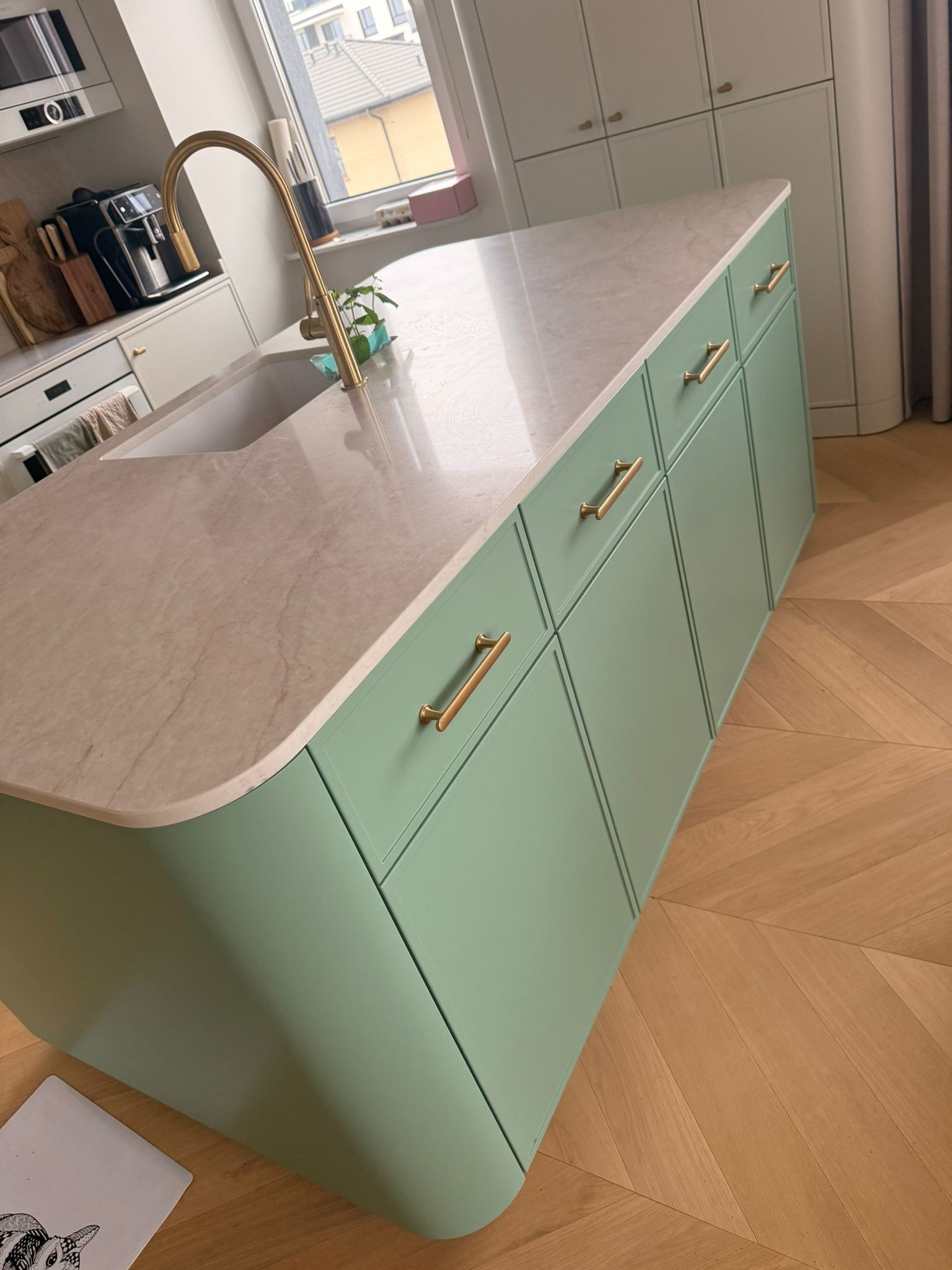 Nowoczesna wyspa kuchenna w pastelowej zieleni z marmurowym blatem i złotymi uchwytami, na tle parkietu w jodełkę. Minimalistyczny design z elementami luksusu.
