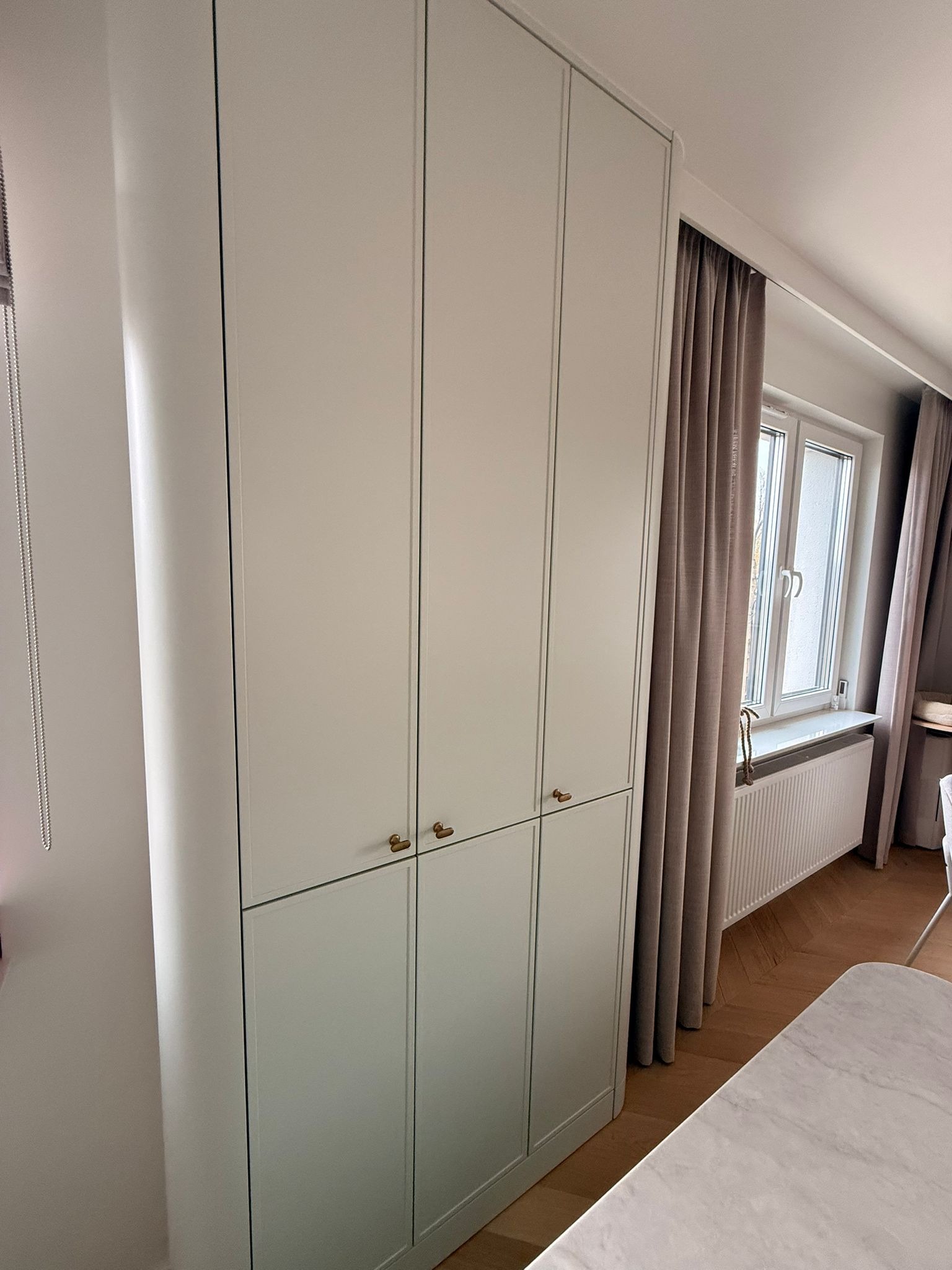 Jasna szafa wnękowa z subtelnymi, złotymi uchwytami, dopasowana do wnętrza w Lublinie. Widok na okno z zasłonami i fragment blatu stołu.