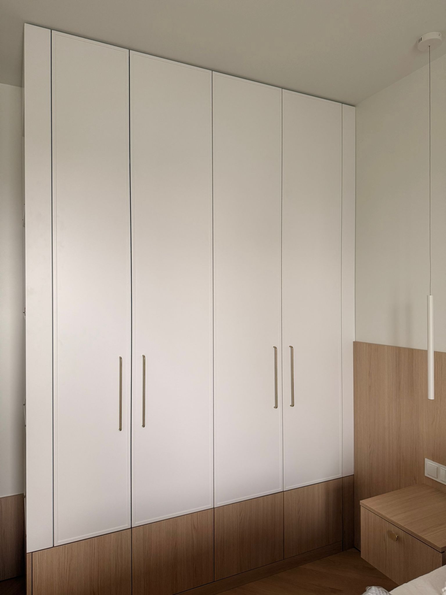 Biała szafa wnękowa z drewnianą podstawą i złotymi uchwytami w sypialni, minimalistyczny design, oświetlenie punktowe.