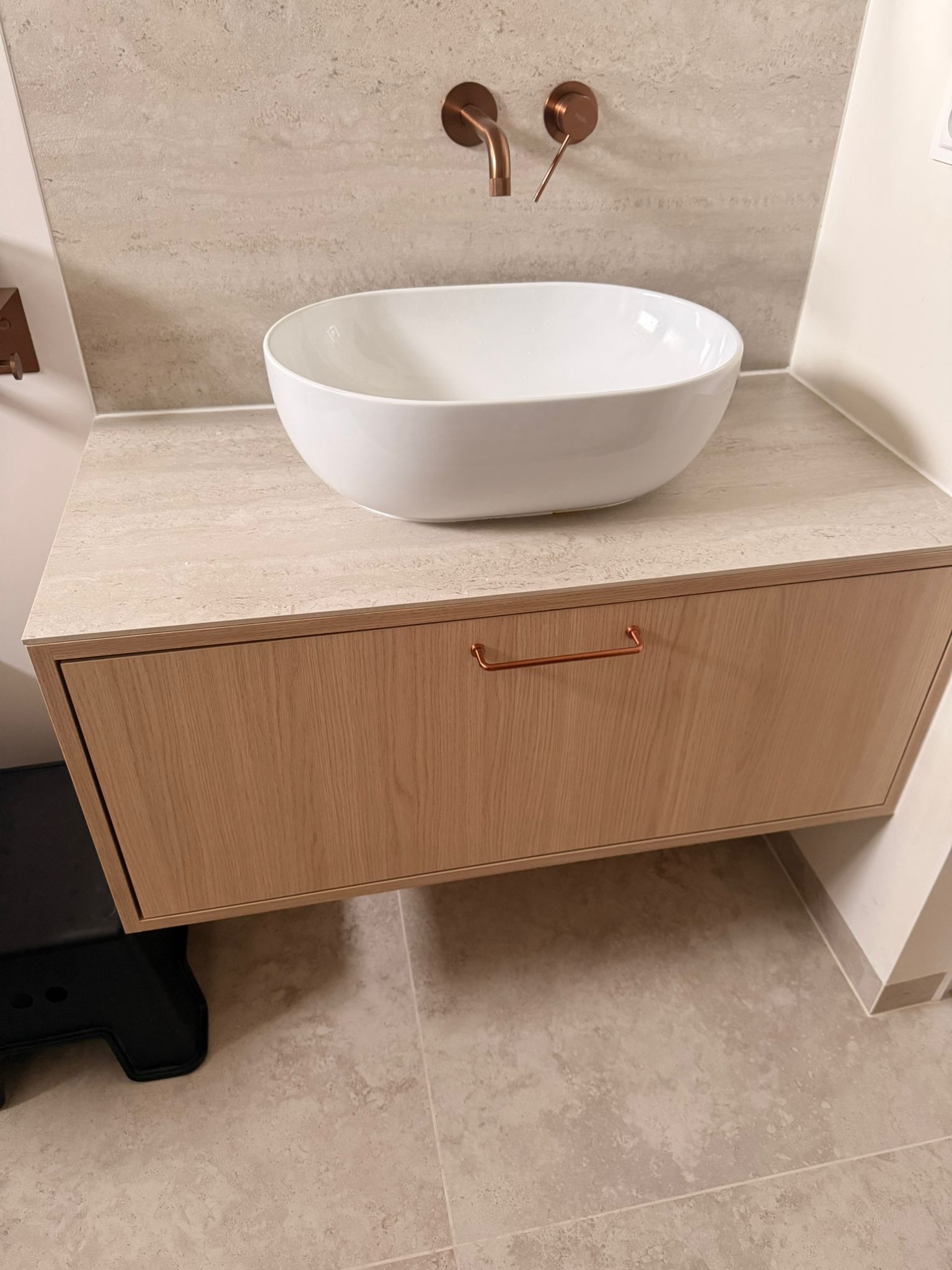 Nowoczesna łazienka: biała, owalna umywalka nablatowa na szafce z jasnego drewna, armatura w kolorze miedzi na tle beżowej ściany z płytek.