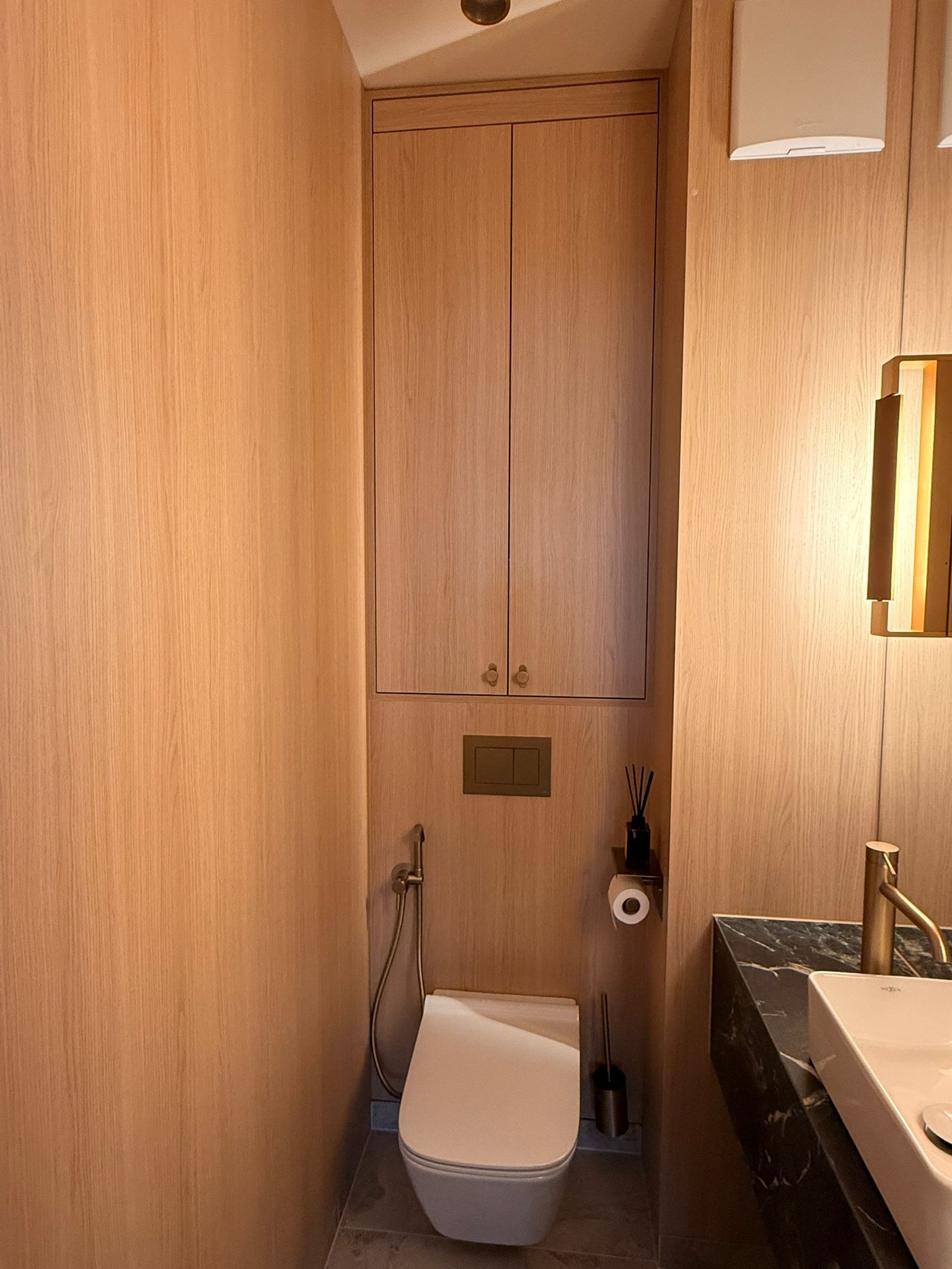 Nowoczesna łazienka z drewnianą szafką nad toaletą, złotą armaturą i blatem z czarnego marmuru. Minimalistyczny design, ciepłe oświetlenie.