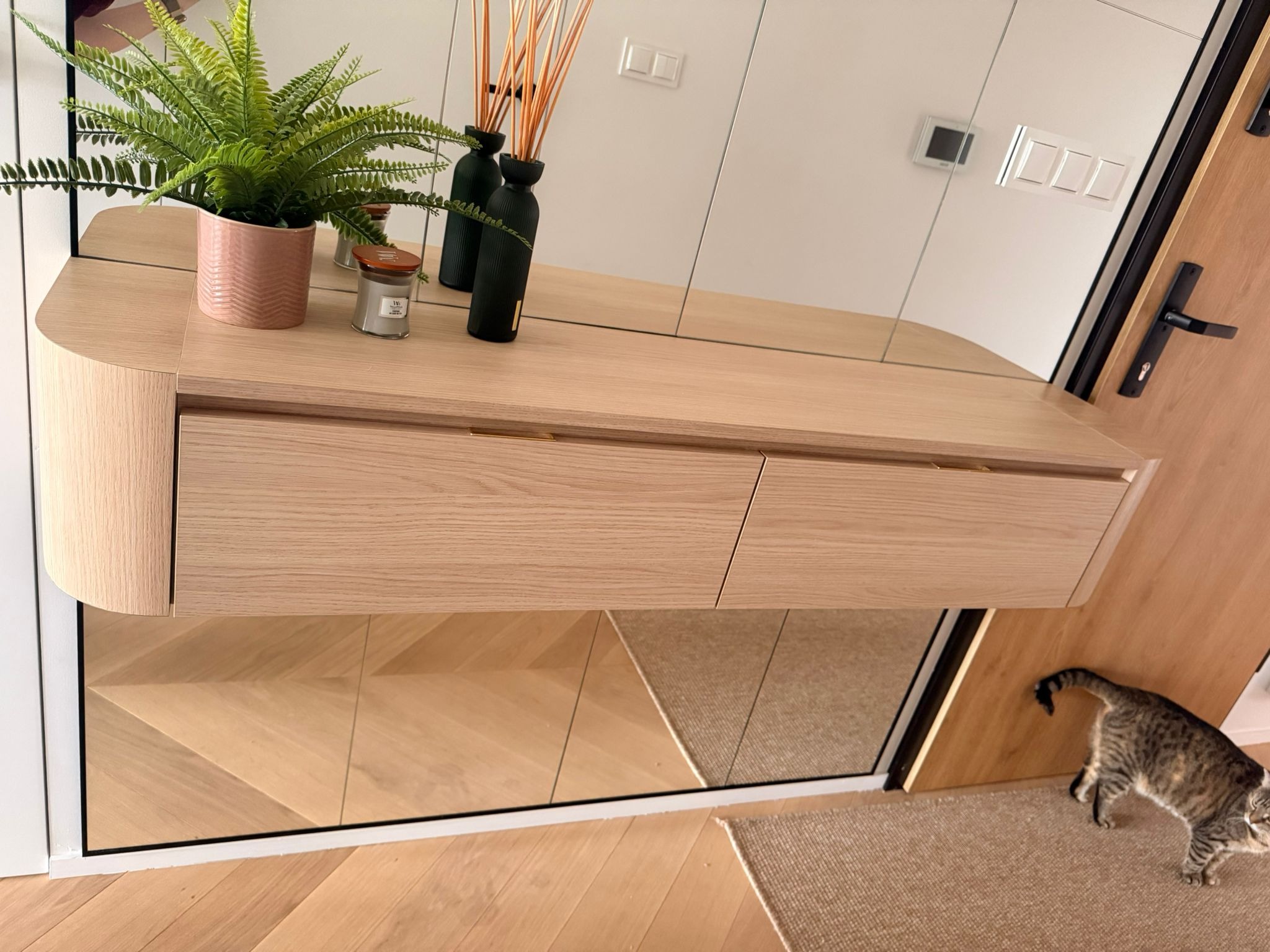 Nowoczesna konsola z lustrem w przedpokoju, drewno w jasnym odcieniu, minimalistyczny design, dekoracje na blacie, kot w tle. Unikalne ujęcie z perspektywy podłogi.