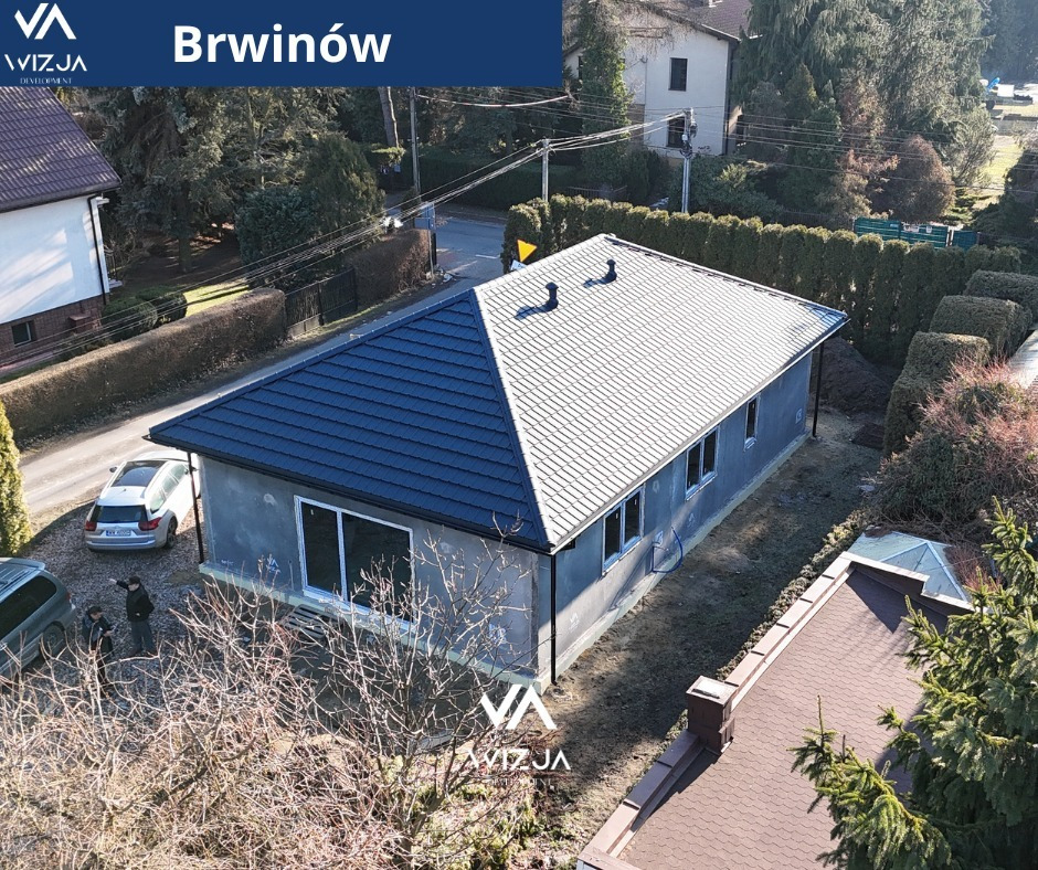 Surowy, parterowy budynek z dwuspadowym dachem w Brwinowie, widziany z góry. Część dachu w kolorze grafitowym, część szara. Dwie osoby i auta na posesji.