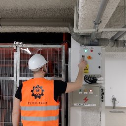 ELITA - TECH Mateusz Pawlak - Pracownik w kamizelce Elita-Tech uruchamia panel sterowania przy metalowej konstrukcji. Widoczne rury pod sufitem i tabliczka ostrzegawcza o ryzyku porażenia prądem.