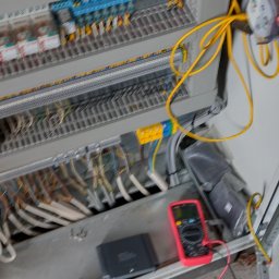 ELITA - TECH Mateusz Pawlak - Otwarta szafa sterownicza z okablowaniem, zabezpieczeniami i multimetrem w kolorze czerwonym, leżącym na podłodze. Widoczne kable i elementy instalacji.