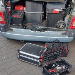 ELITA - TECH Mateusz Pawlak - Otwarty bagażnik auta wypełniony skrzynkami narzędziowymi i pojemnikami, dodatkowe skrzynki na kostce brukowej. Widok z tyłu pojazdu.