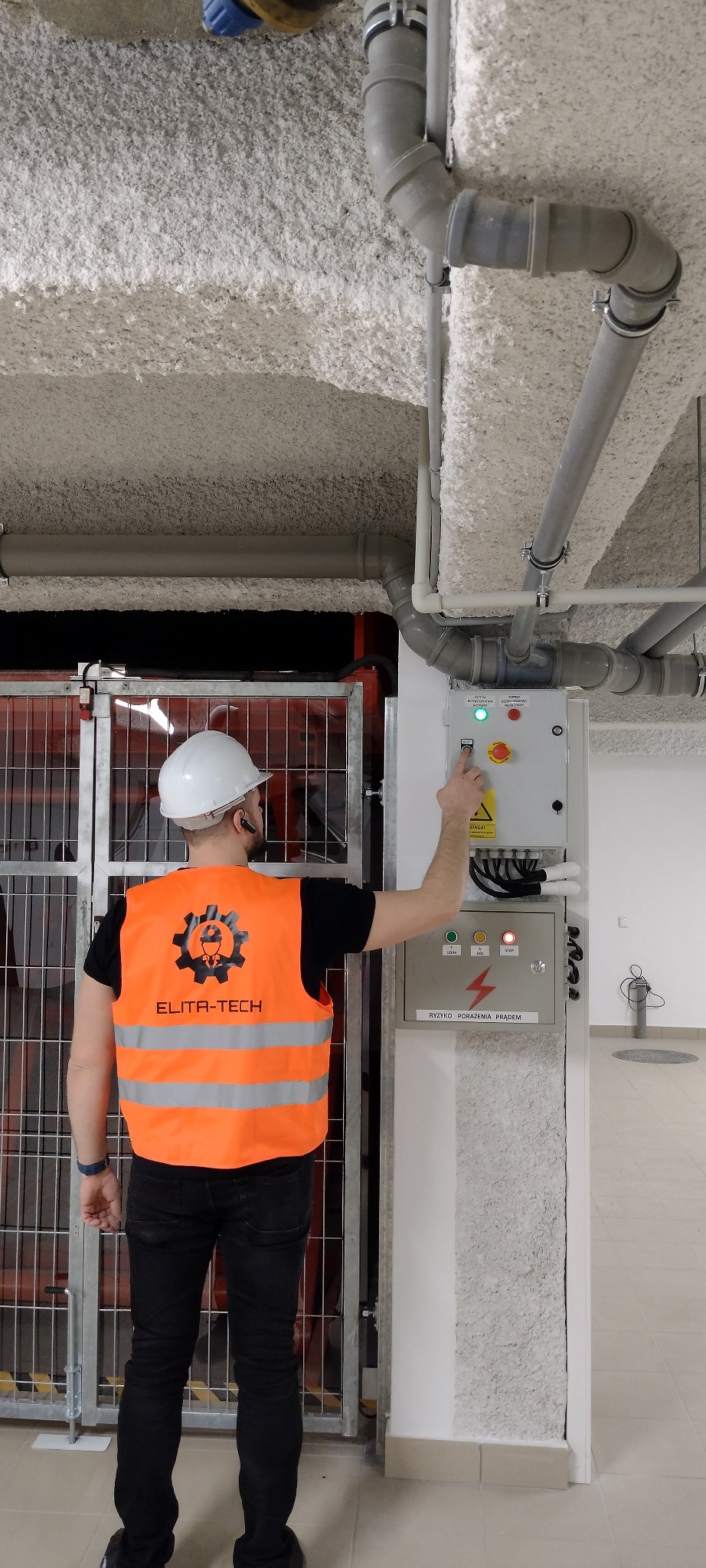 Pracownik w kamizelce Elita-Tech uruchamia panel sterowania przy metalowej konstrukcji. Widoczne rury pod sufitem i tabliczka ostrzegawcza o ryzyku porażenia prądem.