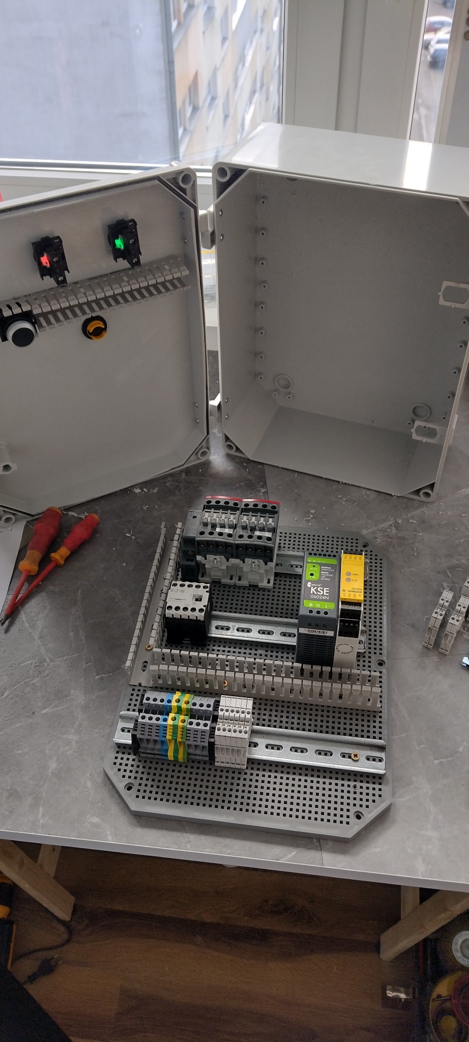 Rozdzielnica elektryczna w trakcie montażu, widoczne elementy sterujące, przewody i narzędzia. Otwarta obudowa rozdzielnicy z widocznymi elementami wewnątrz.