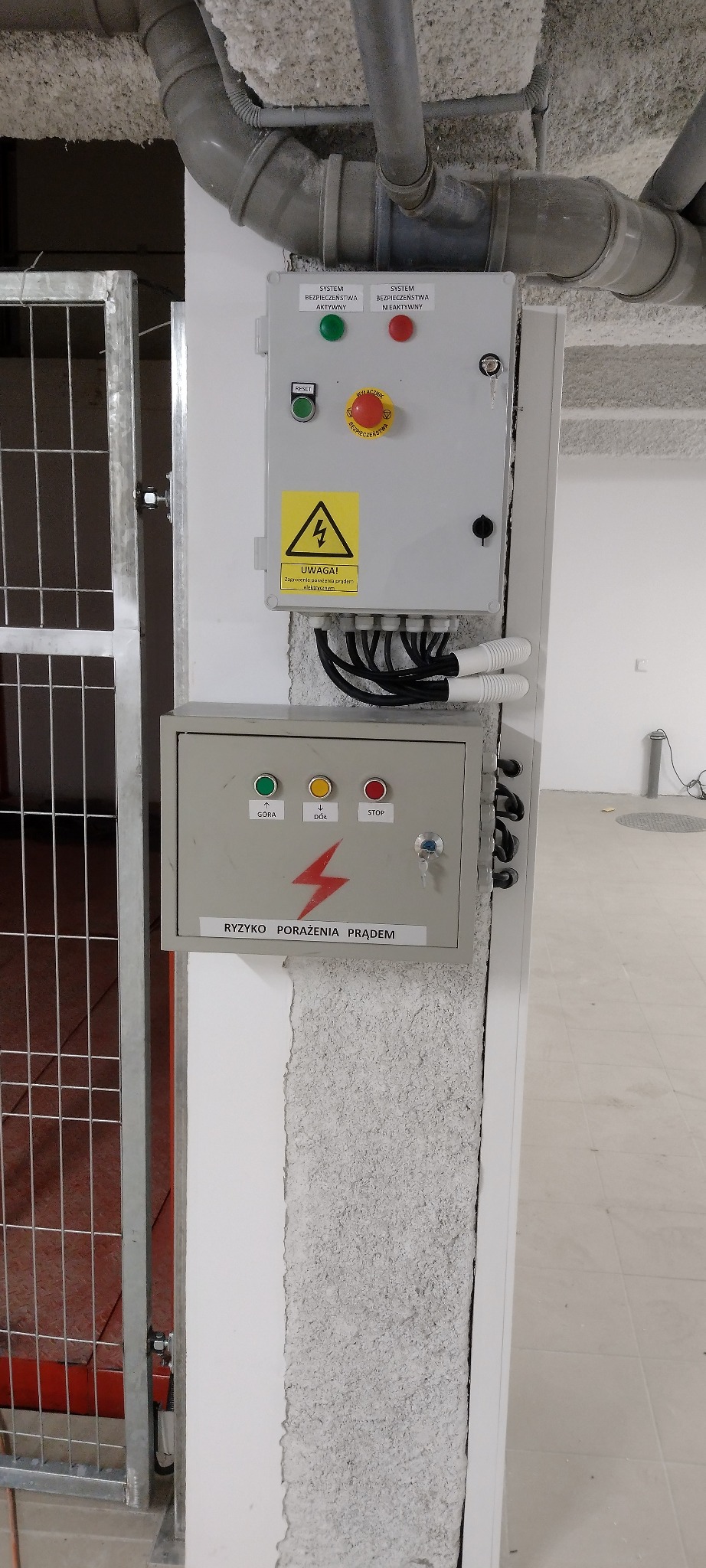 Dwa szare panele sterowania elektrycznego zamontowane na kolumnie, z widocznymi przewodami, przyciskami i ostrzeżeniami o ryzyku porażenia prądem.