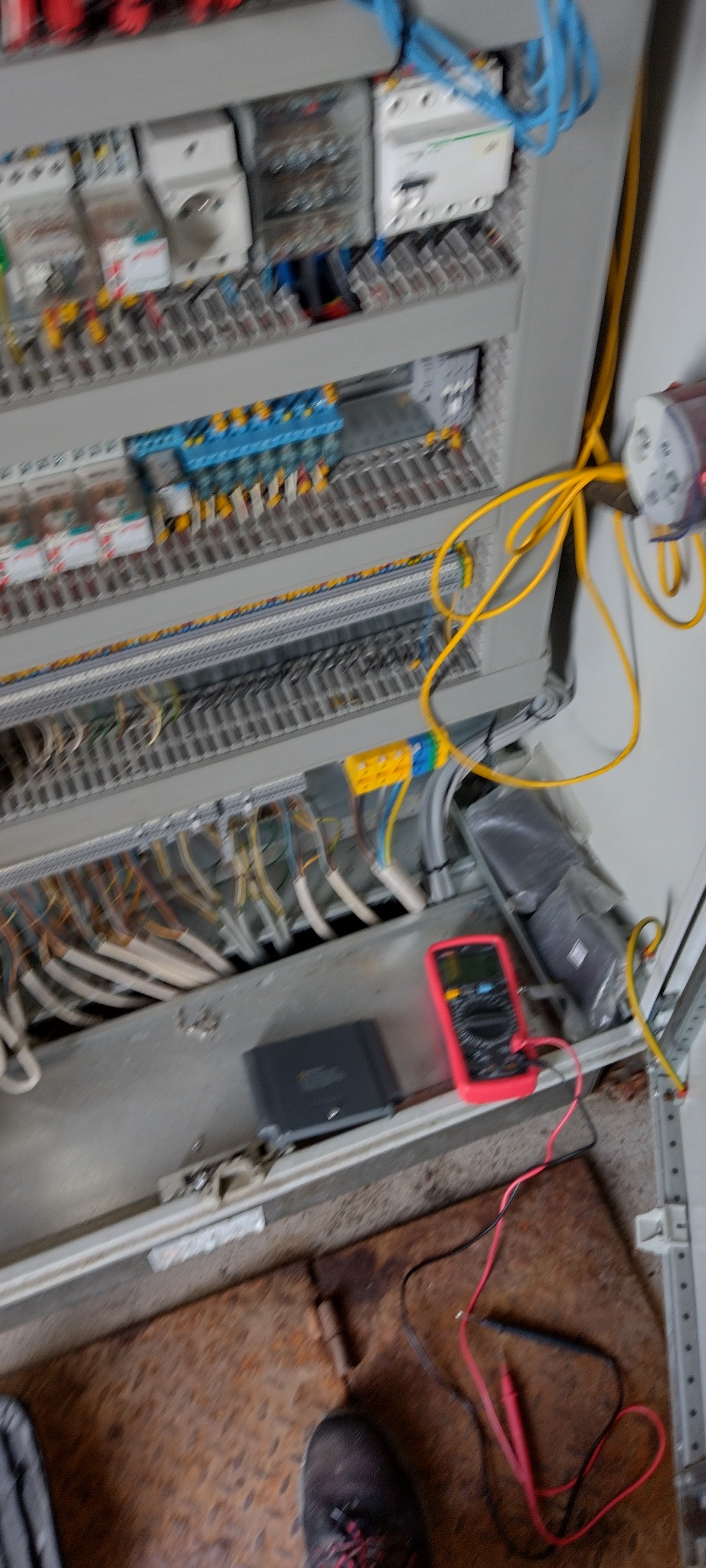 Otwarta szafa sterownicza z okablowaniem, zabezpieczeniami i multimetrem w kolorze czerwonym, leżącym na podłodze. Widoczne kable i elementy instalacji.