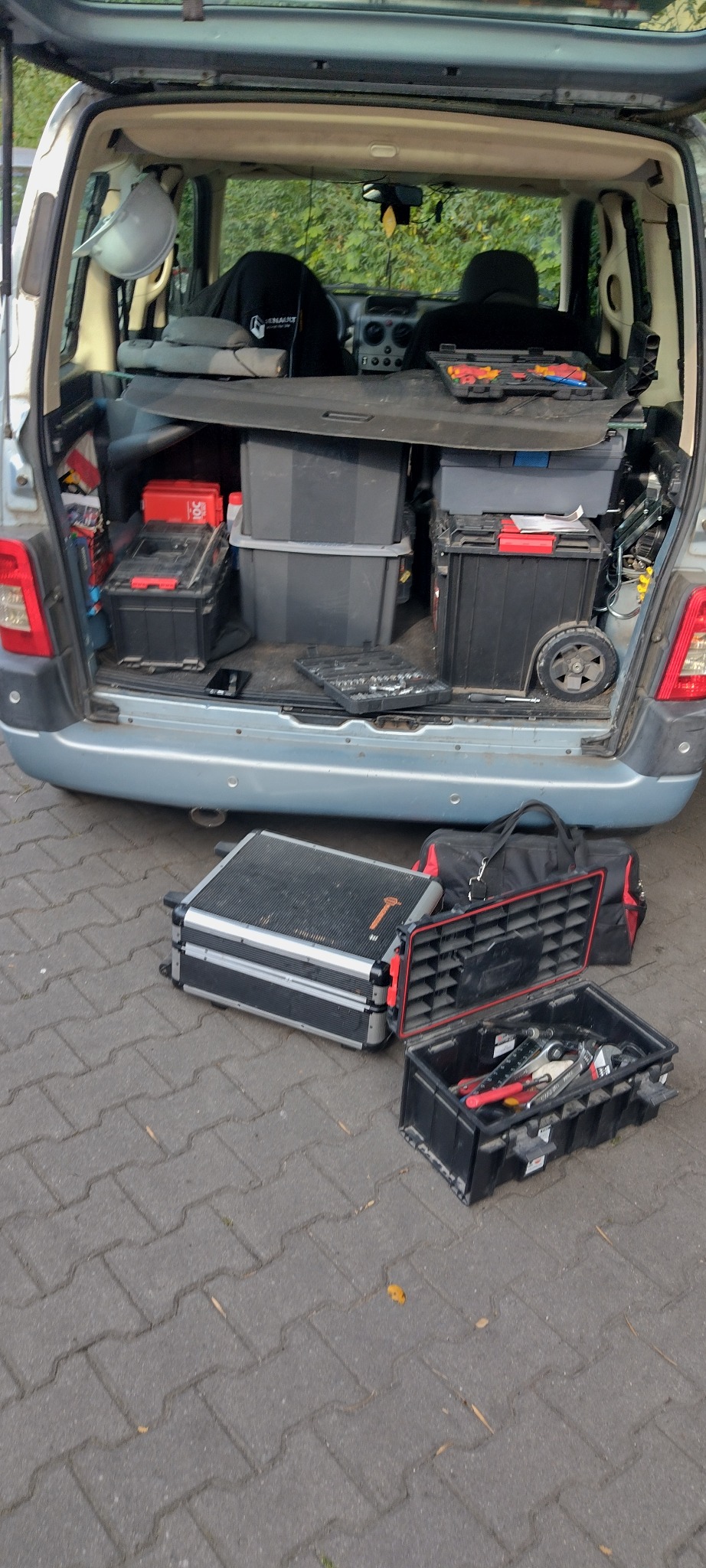 Otwarty bagażnik auta wypełniony skrzynkami narzędziowymi i pojemnikami, dodatkowe skrzynki na kostce brukowej. Widok z tyłu pojazdu.