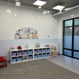 Przedszkole Teddy - Jasne wnętrze sali zabaw z regałami na zabawki, dekoracją ścienną z motywem miasteczka i wiszącymi ozdobami w kształcie chmur. Minimalistyczny design, stonowana kolorystyka.