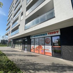 Przedszkole Teddy - Fasada budynku z szyldem 'Przedszkole Teddy' z ofertą zajęć polsko-angielskich i czesnym 0 zł. Widoczne okna z grafikami dzieci i informacjami kontaktowymi.