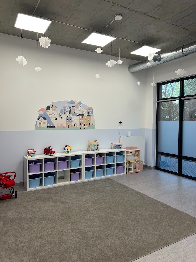 Jasne wnętrze sali zabaw z regałami na zabawki, dekoracją ścienną z motywem miasteczka i wiszącymi ozdobami w kształcie chmur. Minimalistyczny design, stonowana kolorystyka.