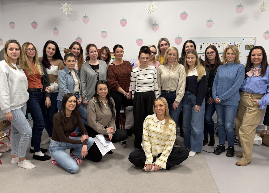 Grupowe zdjęcie uśmiechniętych kobiet, prawdopodobnie pracowniczek żłobka, stojących i klęczących na tle ściany z dekoracjami w kształcie truskawek i śnieżynek.