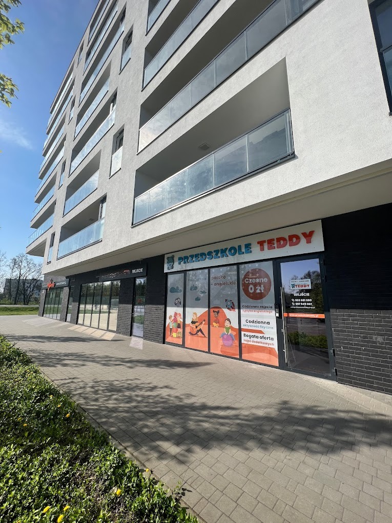 Fasada budynku z szyldem 'Przedszkole Teddy' z ofertą zajęć polsko-angielskich i czesnym 0 zł. Widoczne okna z grafikami dzieci i informacjami kontaktowymi.