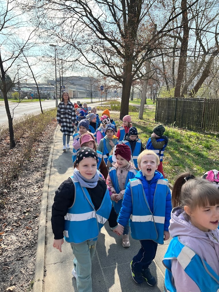 Grupa dzieci w niebieskich kamizelkach idzie chodnikiem w słoneczny dzień, prowadzona przez opiekunkę w czarno-białej kracie. W tle drzewa i budynki.
