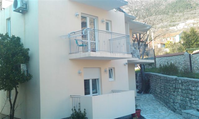 Apartamenty w jasnych kolorach z balkonami z metalowymi barierkami, widok na kamienny murek i zbocze góry w tle.