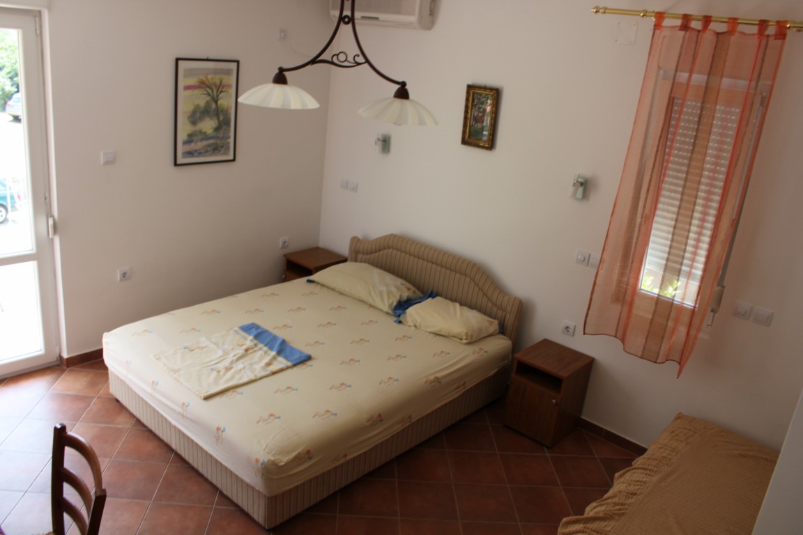 Pokój z beżowym łóżkiem, wzorzystą pościelą i niebieskim ręcznikiem, oświetlony wiszącą lampą z trzema kloszami, z pomarańczową zasłoną w oknie i brązowymi płytkami na podłodze.