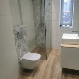 Usługi glazurnicze - Nowoczesna łazienka po remoncie: białe płytki, drewnopodobna podłoga, kabina prysznicowa ze wzorem, podwieszana toaleta i umywalka nablatowa. Widok na okno.