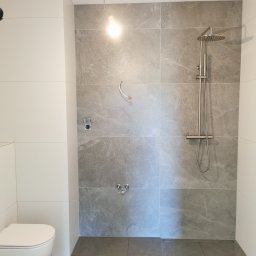 Usługi glazurnicze - Nowoczesna łazienka w Krotoszynie z szarymi płytkami imitującymi kamień, białą armaturą i deszczownicą. Minimalistyczny design, surowy styl.