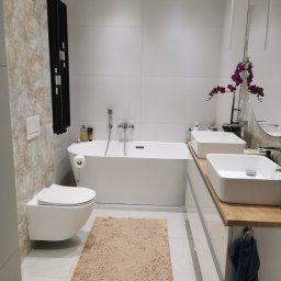 Usługi glazurnicze - Nowoczesna łazienka z wolnostojącą wanną, podwieszaną toaletą i dwiema umywalkami nablatowymi. Uwagę zwraca dekoracyjna ściana z efektem kamienia i minimalistyczne oświetlenie.