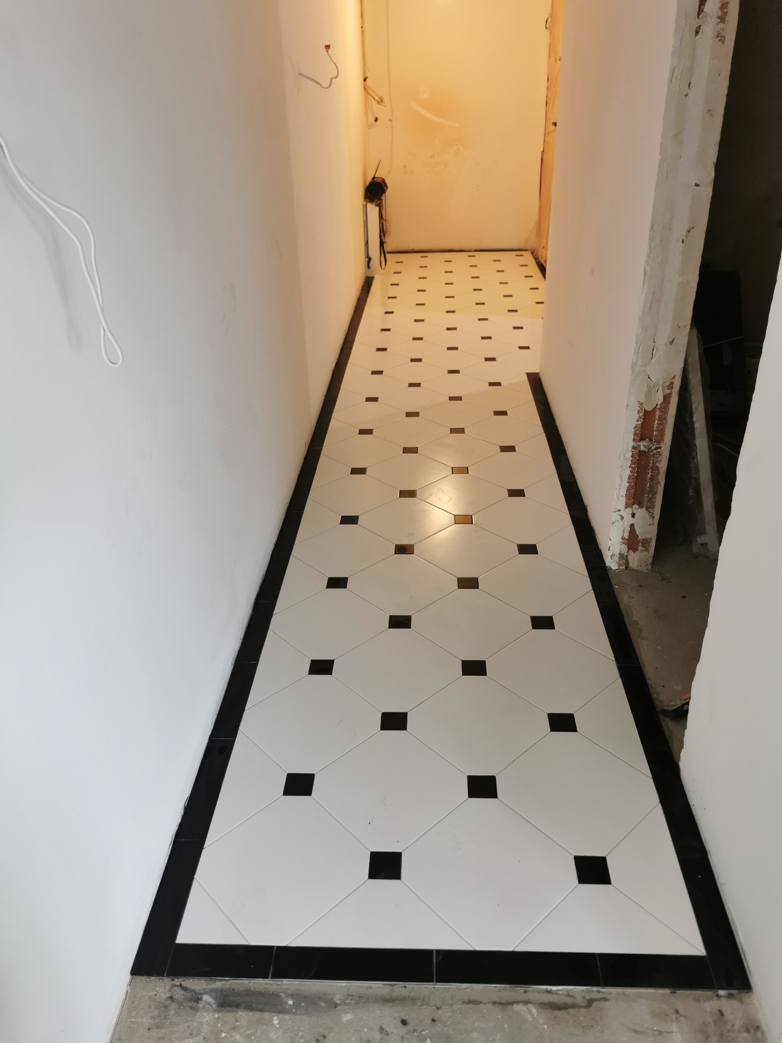 Ukończone układanie białych płytek z czarnymi akcentami w wąskim korytarzu. Widoczna czarna listwa przypodłogowa i fragmenty surowych ścian.
