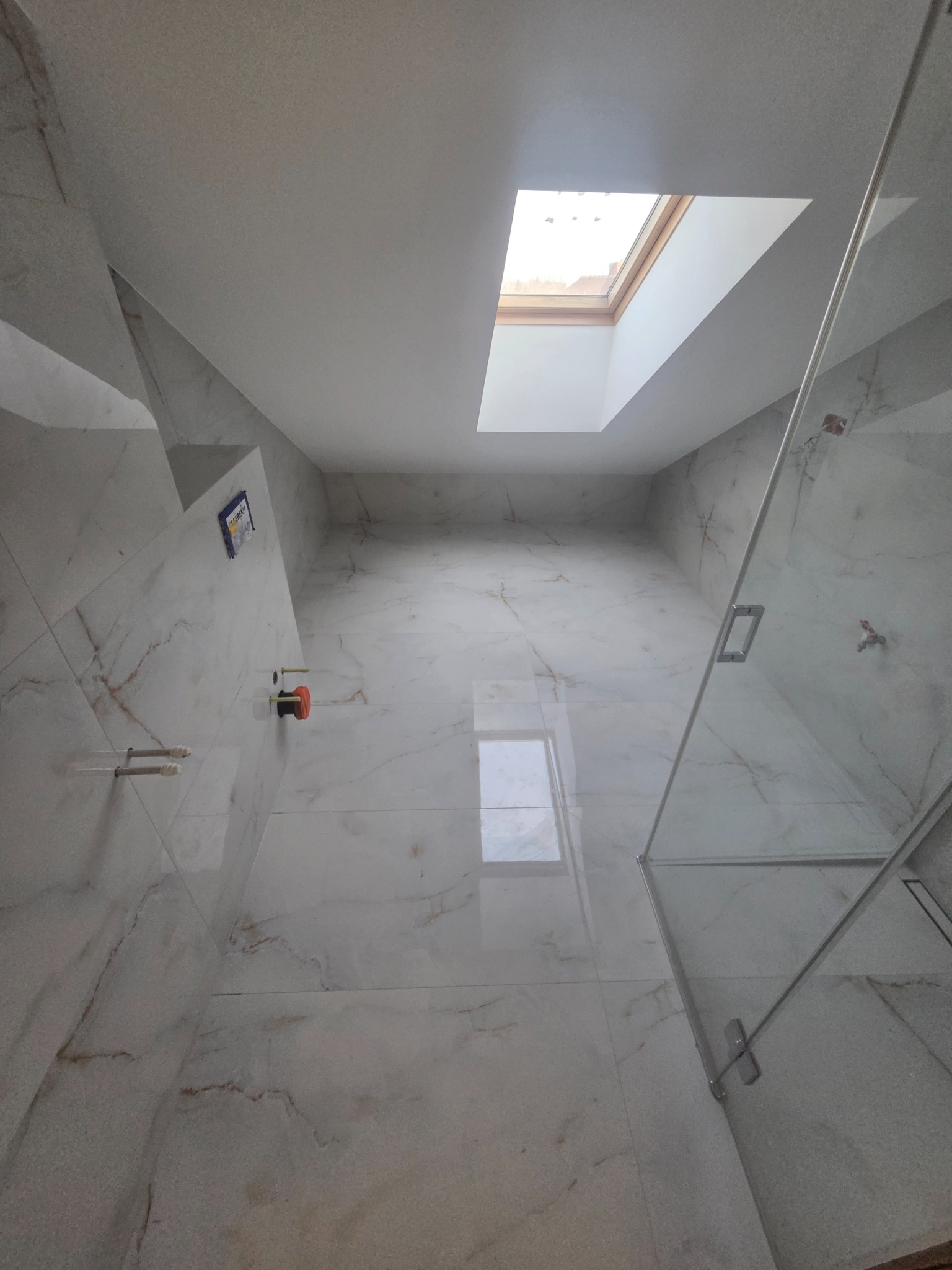 Jasna łazienka na poddaszu z marmurowymi płytkami, kabiną prysznicową ze szklanymi drzwiami i oknem dachowym w suficie. Widok z dołu, podkreślający przestrzeń i wykończenie.